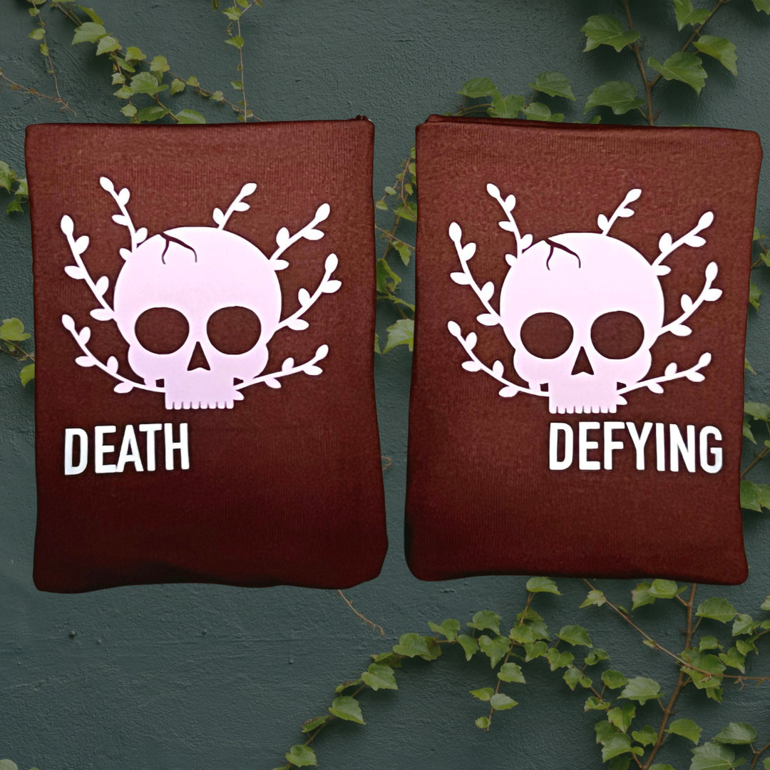Two rectangular dark brown DRYbands, best wristbands for gymnasts, with a light pink skull and leaf graphic, one labeled 'DEATH' and the other 'DEFYING', displayed against a dark green textured wall with ivy vines.