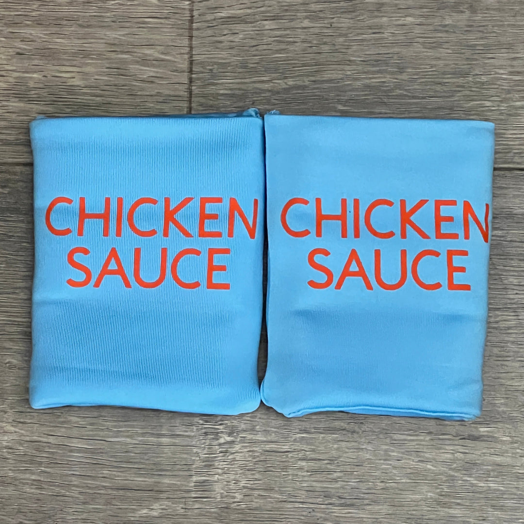 Two light blue DRYbands, best wristbands for gymnasts, with 'CHICKEN SAUCE' printed in orange, displayed side-by-side on a gray wood-grain surface.