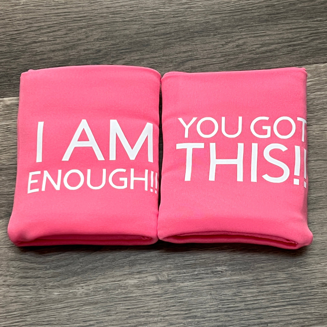 Two pink DRYbands, best wristbands for gymnasts with white text, placed side by side on a gray wood surface. The left one reads 'I AM ENOUGH!!' and the right one reads 'YOU GOT THIS!!'.