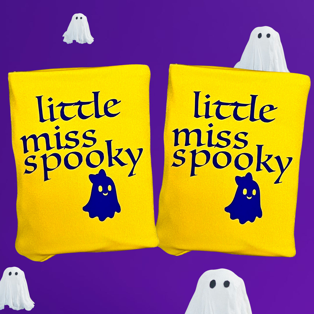 Two bright yellow DRYbands gymnastics wristbands with the phrase 'little miss spooky' in dark purple lettering and a small purple ghost graphic, shown on a purple background with white ghost illustrations around them.