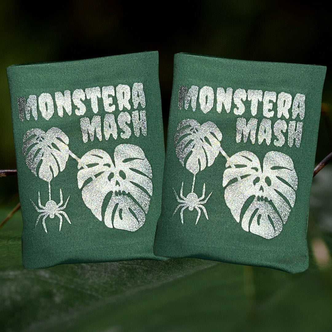 Two sage green DRYbands, best wristbands for gymnasts with silver glitter text reading 'MONSTERA MASH' and silver monster graphics, shown outdoors against a blurred green background.