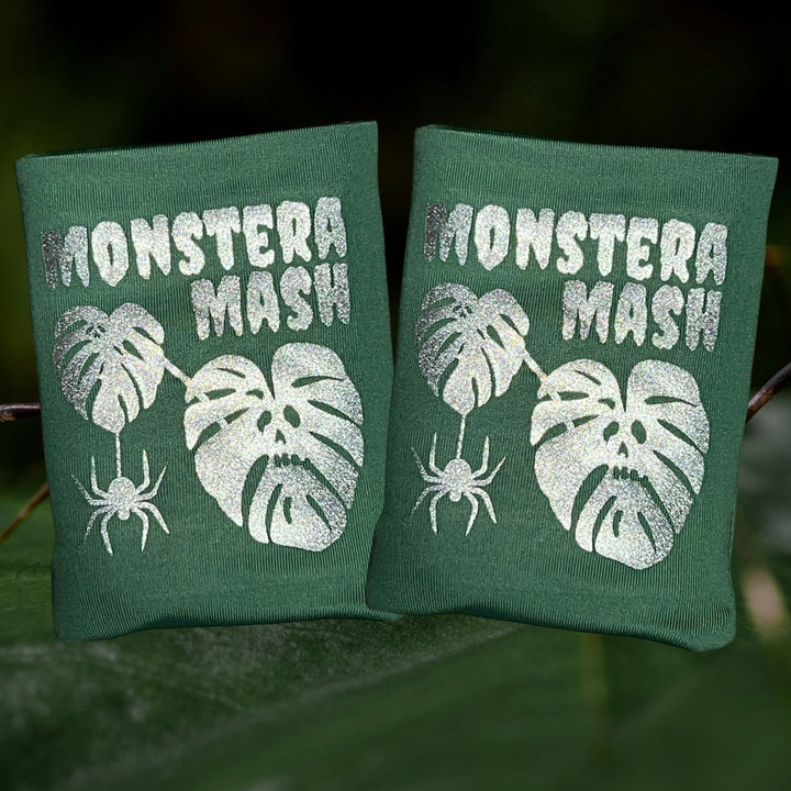 Two sage green DRYbands, best wristbands for gymnasts with silver glitter text reading 'MONSTERA MASH' and silver monster graphics, shown outdoors against a blurred green background.
