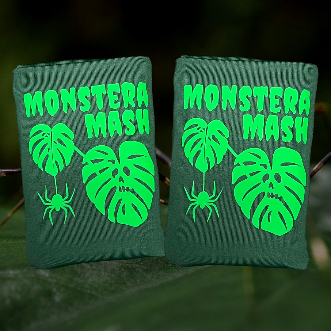Two dark green DRYbands, best wristbands for gymnasts, with bright green 'MONSTERA MASH' text, monstera leaf graphics, and spider illustrations, displayed outdoors on a leaf with a blurred green background.